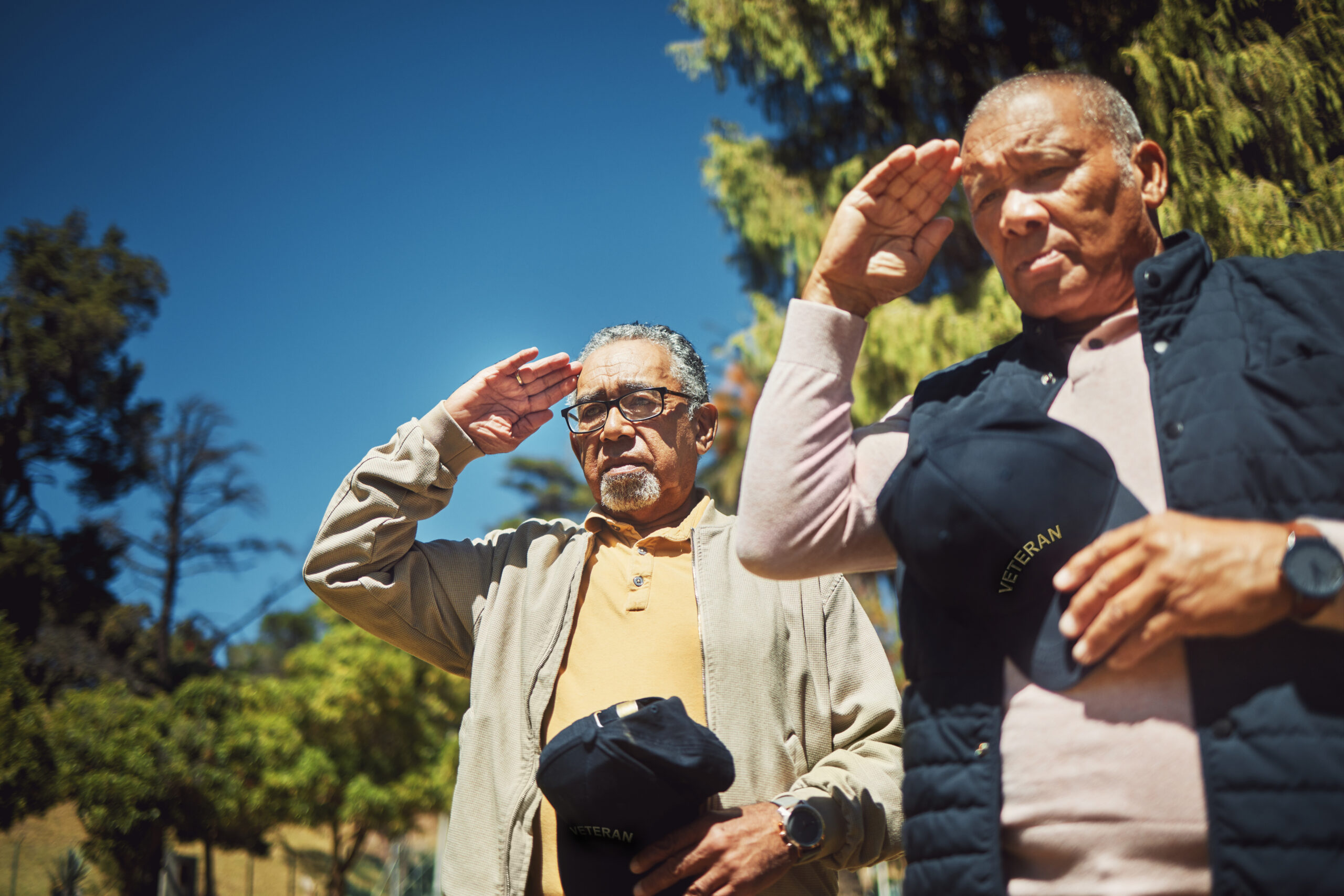 Two elderly Veterans saluting with their right hands on their forehead. Each man has a hat in their left hand. Trees and clear skies are in the background.