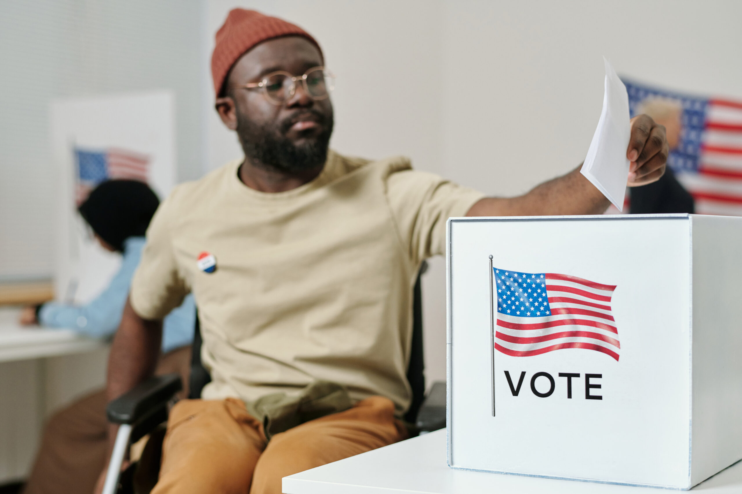 Box for ballot papers on desk and young African American man with disability sitting in wheelchair and making his choice.
