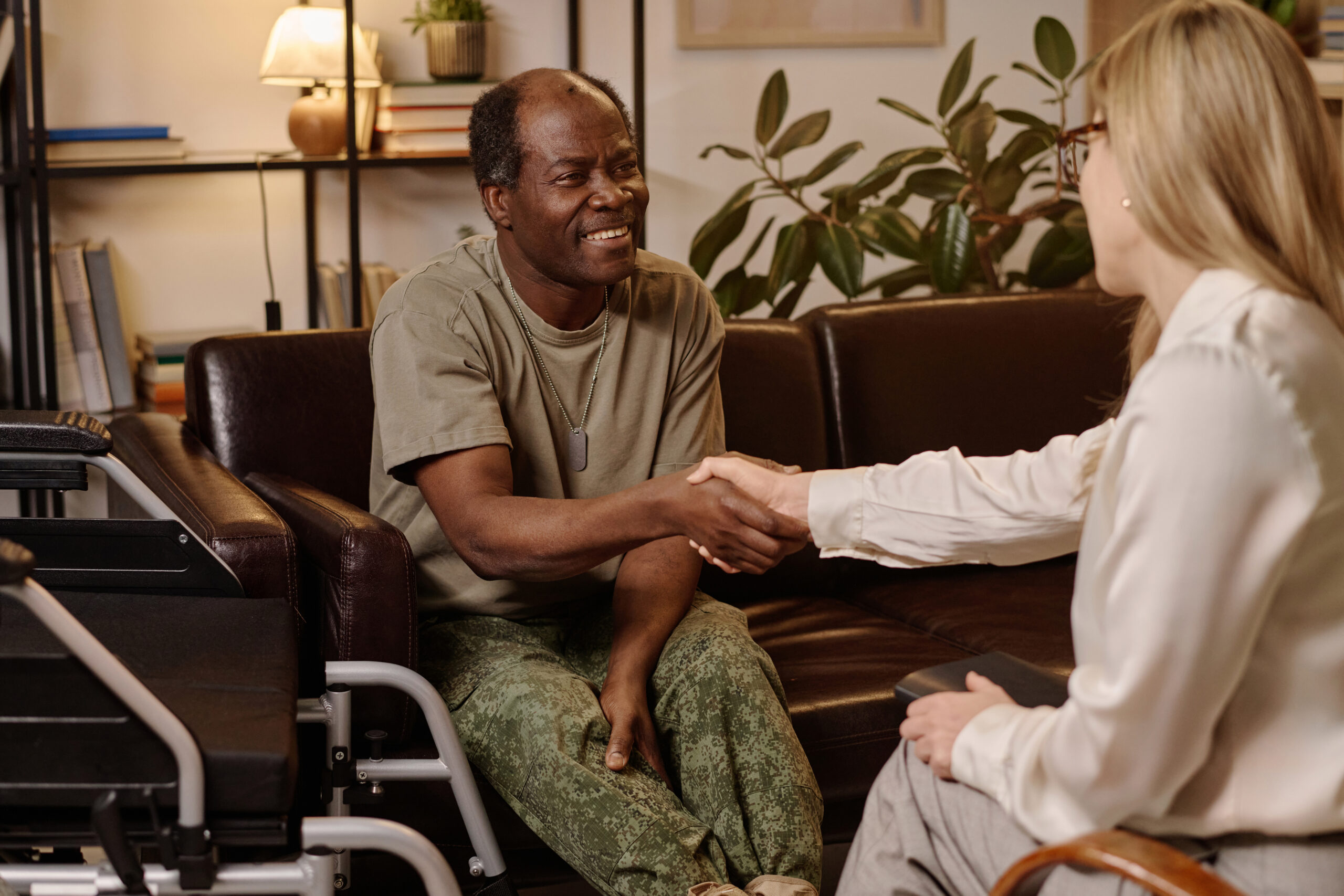 Joyful black man with disability shaking hands with female psychologist sitting on sofa at office