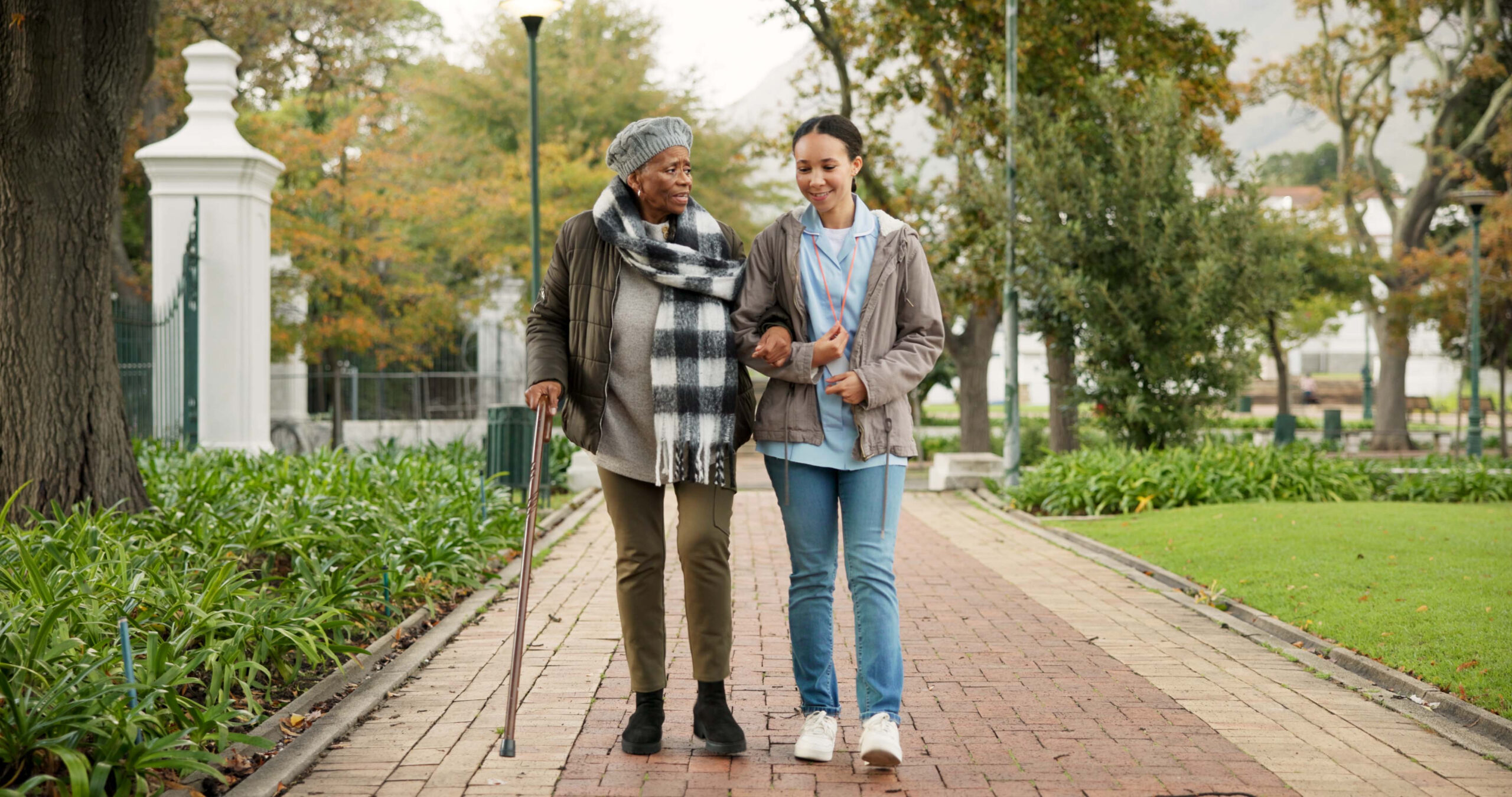 Caregiver (right) with aging woman (left) walking in the park down a brick path.